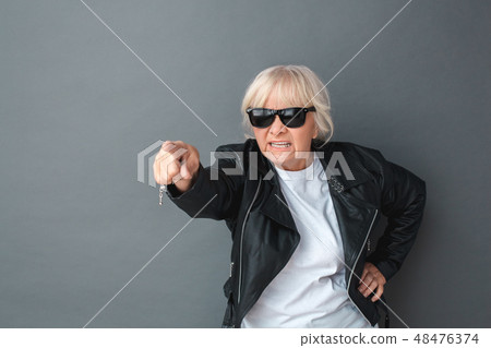 Senior woman in leather jacket and sunglasses studio standing isolated on gray pointing camera Senior woman in leather jacket and sunglasses studio standing isolated on gray pointing camera 48476374