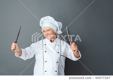 Senior woman chef studio standing isolated on gray playing with knives hands up smiling Senior woman chef studio standing isolated on gray playing with knives hands up smiling 48477267