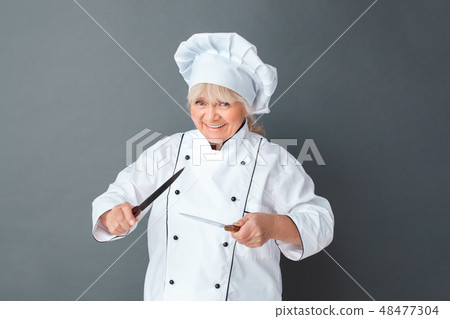Senior woman chef studio standing isolated on gray with knives frightening playful 48477304