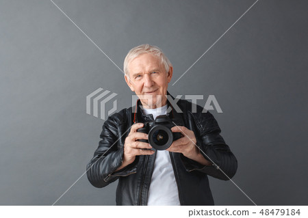 Senior man in leather jacket standing isolated on gray holding camera happy Senior man in leather jacket standing isolated on gray holding camera happy 48479184