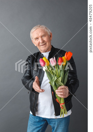 Senior man in leather jacket standing isolated on gray giving bouquet of tulips to camera cheerful 48479205