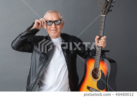 Senior man in leather jacket and sunglasses standing isolated on gray holding guitar touching 48480012