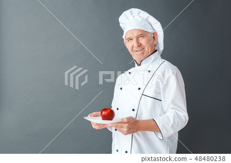 Senior chef studio standing isolated on gray with apple on dish looking camera cheerful 48480238