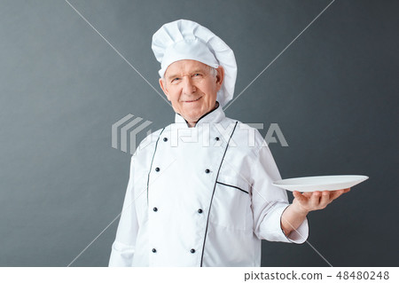 Senior chef studio standing isolated on gray serving plate looking camera joyful Senior chef studio standing isolated on gray serving plate looking camera joyful 48480248