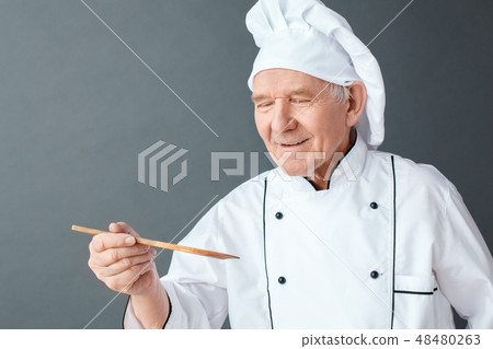 Senior chef studio standing isolated on gray tasting dish with spatula smiling joyful close-up Senior chef studio standing isolated on gray tasting dish with spatula smiling joyful close-up 48480263