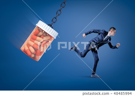 A side view of a businessman running from a huge full pills jar swinging on a chain on a blue A side view of a businessman running from a huge full pills jar swinging on a chain on a blue 48485994