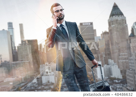 In a hurry. Portrait of young confident businessman in suit pulling luggage and talking by phone 48486632