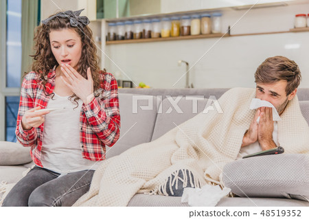 A man with a cold sitting on a sofa in a blanket blanket. Checking the temperature. The wife is A man with a cold sitting on a sofa in a blanket blanket. Checking the temperature. The wife is 48519352