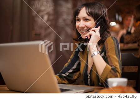 Pretty young woman smiling while talking on the phone and using her laptop in a coffee shop Pretty young woman smiling while talking on the phone and using her laptop in a coffee shop 48542196