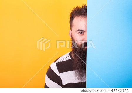 Handsome bearded hipster hiding behind a blue panel over yellow background Handsome bearded hipster hiding behind a blue panel over yellow background 48542995