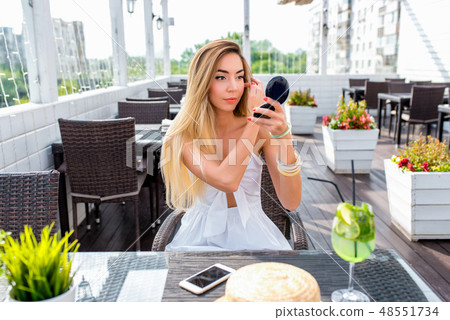 A young and beautiful girl in summer in a cafe at a table, paints make-up, powder mirror. At the 48551734