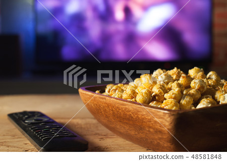 A wooden bowl of popcorn and remote control. 48581848