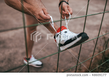 Close-up of a man's leg tying shoelaces on a shoe, in summer in the city about a fence or fence 48609177
