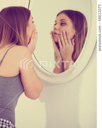 Young female touching face, using mirror in bedroom Young female touching face, using mirror in bedroom 48612075
