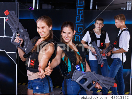 Two smiling girls posing with plastic laser pistols and two guys in background in laser tag room 48615522