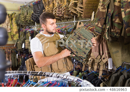 Young guy choosing flak jacket in military shop Young guy choosing flak jacket in military shop 48616963