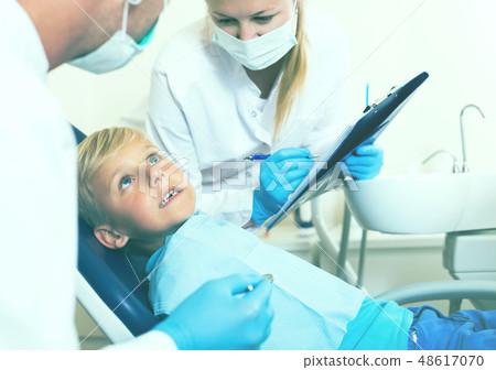 Dentist with woman assistant are diagnosticating to young patient which is sitting Dentist with woman assistant are diagnosticating to young patient which is sitting 48617070