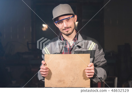 Portrait of a young carpenter in overalls and goggles with a mock up board holding a blank sign in 48632529