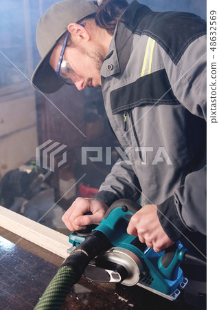 Young carpenter with a beard working with an electric plane with suction of sawdust. Leveling and 48632569