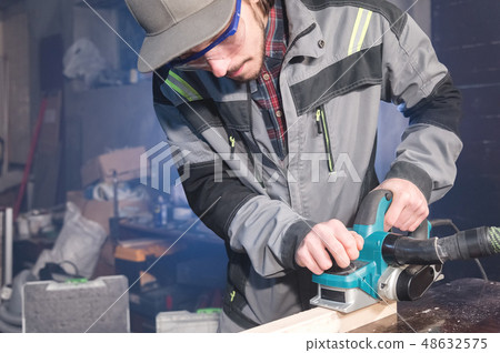 Young carpenter with a beard working with an electric plane with suction of sawdust. Leveling and Young carpenter with a beard working with an electric plane with suction of sawdust. Leveling and 48632575