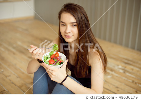 Sports beautiful girl sitting on the floor and eating a salad from fresh vegetables and greens Sports beautiful girl sitting on the floor and eating a salad from fresh vegetables and greens 48636179