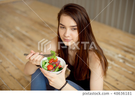 Sports beautiful girl sitting on the floor and eating a salad from fresh vegetables and greens Sports beautiful girl sitting on the floor and eating a salad from fresh vegetables and greens 48636181