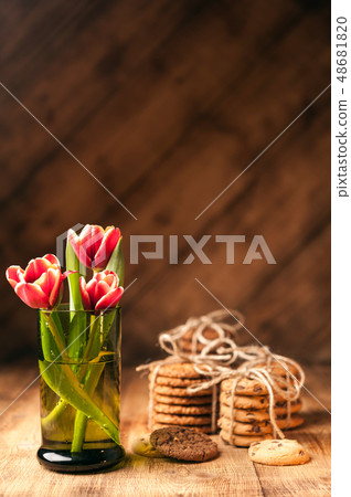 Simple rustic still life of red tulips in a green glass and stacks various cookies on wooden table. Simple rustic still life of red tulips in a green glass and stacks various cookies on wooden table. 48681820