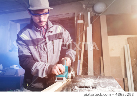 Young carpenter with a beard working with an electric plane with suction of sawdust. Leveling and 48683298