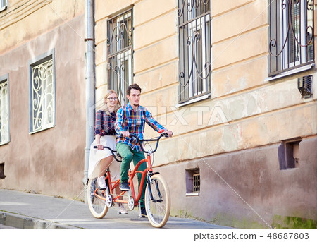 Young tourist couple, handsome man and pretty blond woman riding tandem bicycle along city street. 48687803