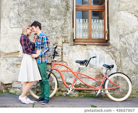 Young tourist couple, handsome man and pretty blond woman riding tandem bicycle along city street. 48687807