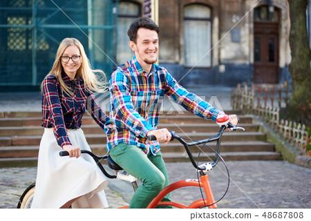 Young tourist couple, handsome man and pretty blond woman riding tandem bicycle along city street. 48687808