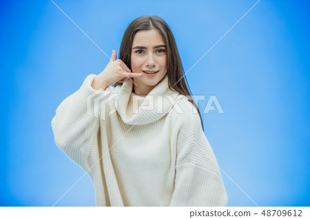 Beautiful young girl on a blue background. The girl shows a phone call with a gesture. Dressed in a Beautiful young girl on a blue background. The girl shows a phone call with a gesture. Dressed in a 48709612