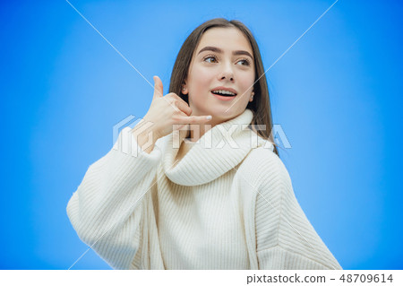 Beautiful young girl on a blue background. The girl shows a phone call with a gesture. Dressed in a Beautiful young girl on a blue background. The girl shows a phone call with a gesture. Dressed in a 48709614