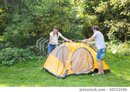 Young happy couple are preparing for camping. They're installing a tent on a suitable place in a Young happy couple are preparing for camping. They're installing a tent on a suitable place in a 48722496