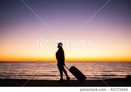 Traveling, beach and holiday concept - Female silhouette walking along ocean coast pulling suitcase Traveling, beach and holiday concept - Female silhouette walking along ocean coast pulling suitcase 48725669