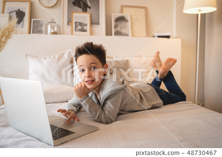 A little boy sitting on a big bed with a laptop book and looks into the camera. A little boy sitting on a big bed with a laptop book and looks into the camera. 48730467