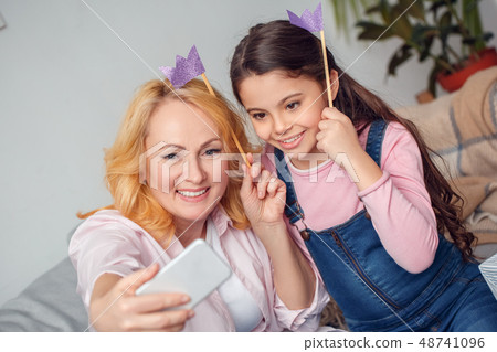 Granddaughter and grandmother at home celebration sitting taking selfie pictures with paper crowns 48741096
