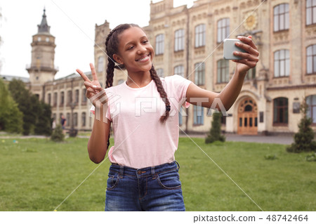 Young schoolgirl standing at school yard taking selfie on smartphone showing peace sign smiling Young schoolgirl standing at school yard taking selfie on smartphone showing peace sign smiling 48742464