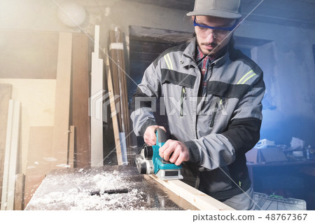 Young carpenter with a beard working with an electric plane with suction of sawdust. Leveling and 48767367