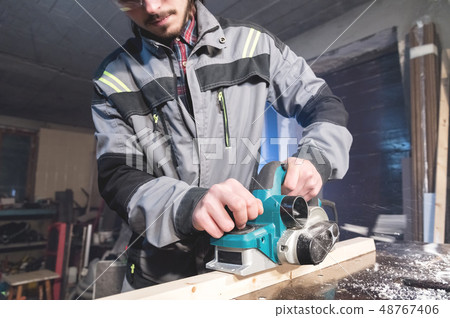 Young carpenter with a beard working with an electric plane with suction of sawdust. Leveling and Young carpenter with a beard working with an electric plane with suction of sawdust. Leveling and 48767406