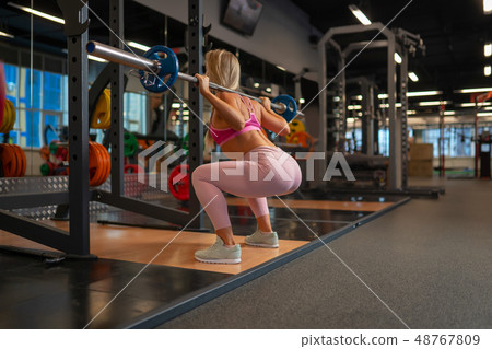 Photo from back of blonde girl crouching with barbell on her shoulders in gym. Photo from back of blonde girl crouching with barbell on her shoulders in gym. 48767809