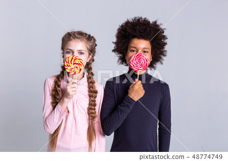 Cute good-looking kids holding sweets and covering half of the face Cute good-looking kids holding sweets and covering half of the face 48774749