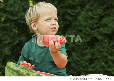 Funny little toddler boy with blond hairs eating watermelon in summer garden. Kid tasting healthy 48809339