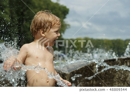 Funny baby boy trying to cauch water stream in fountain. Cute toddler playing in the city fountain Funny baby boy trying to cauch water stream in fountain. Cute toddler playing in the city fountain 48809344