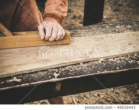 Carpenter in Medieval Cotton Clothes Working With a Wood by Plane. Man Manually Hews a Wooden Board 48816441