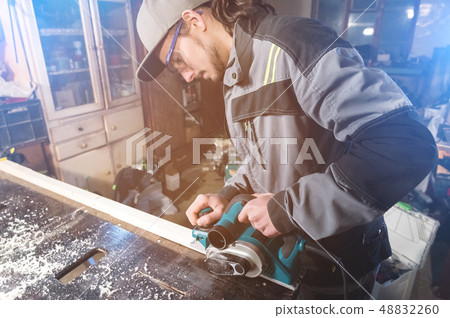 Young carpenter with a beard working with an electric plane with suction of sawdust. Leveling and Young carpenter with a beard working with an electric plane with suction of sawdust. Leveling and 48832260