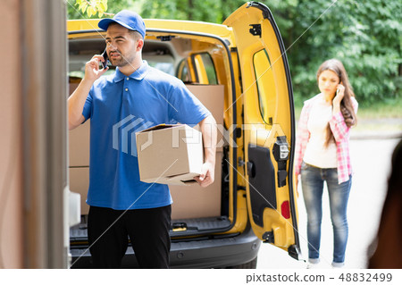Delivery man wearing blue uniform holding cardboard box and talking on phone. Open van in background Delivery man wearing blue uniform holding cardboard box and talking on phone. Open van in background 48832499