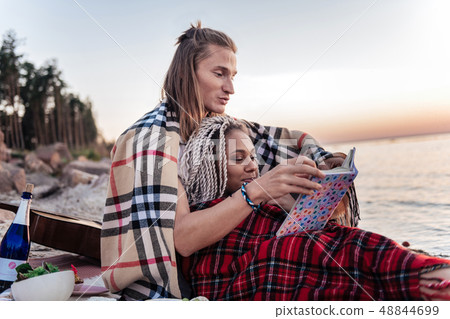Loving cute couple with plaids on shoulders reading book after picnic Loving cute couple with plaids on shoulders reading book after picnic 48844699