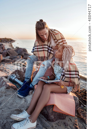 Loving couple sitting with plaid on their shoulders reading book near river Loving couple sitting with plaid on their shoulders reading book near river 48844717