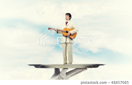 Businessman on metal tray playing acoustic guitar against blue sky background 48857085